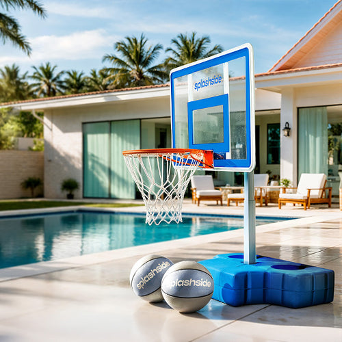 Portable basketball hoop and two balls on a patio near a pool with a house and palm trees in the background.