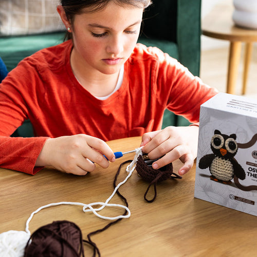 Child in red shirt crocheting with yarn and a crochet owl toy kit on a table.