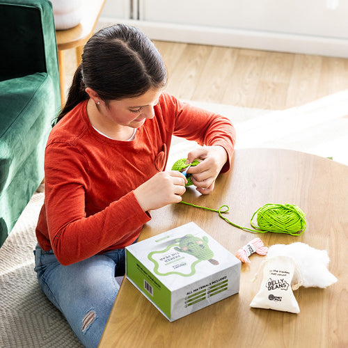 Person sitting on a couch making something with green yarn and a box on a wooden floor.