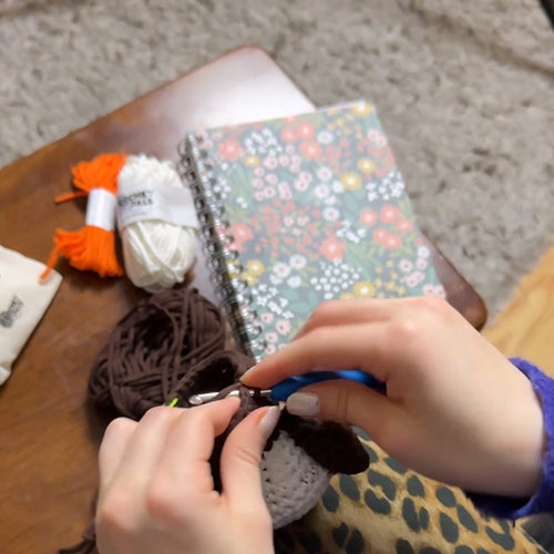 Person cutting yarn with scissors over a notebook and yarn balls on a wooden surface.