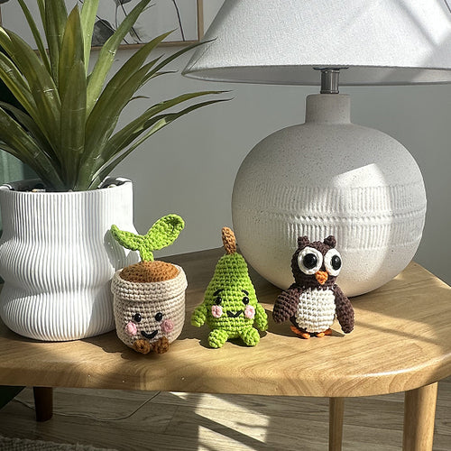 Three small crochet characters on a wooden table with a lamp and plant in the background.

